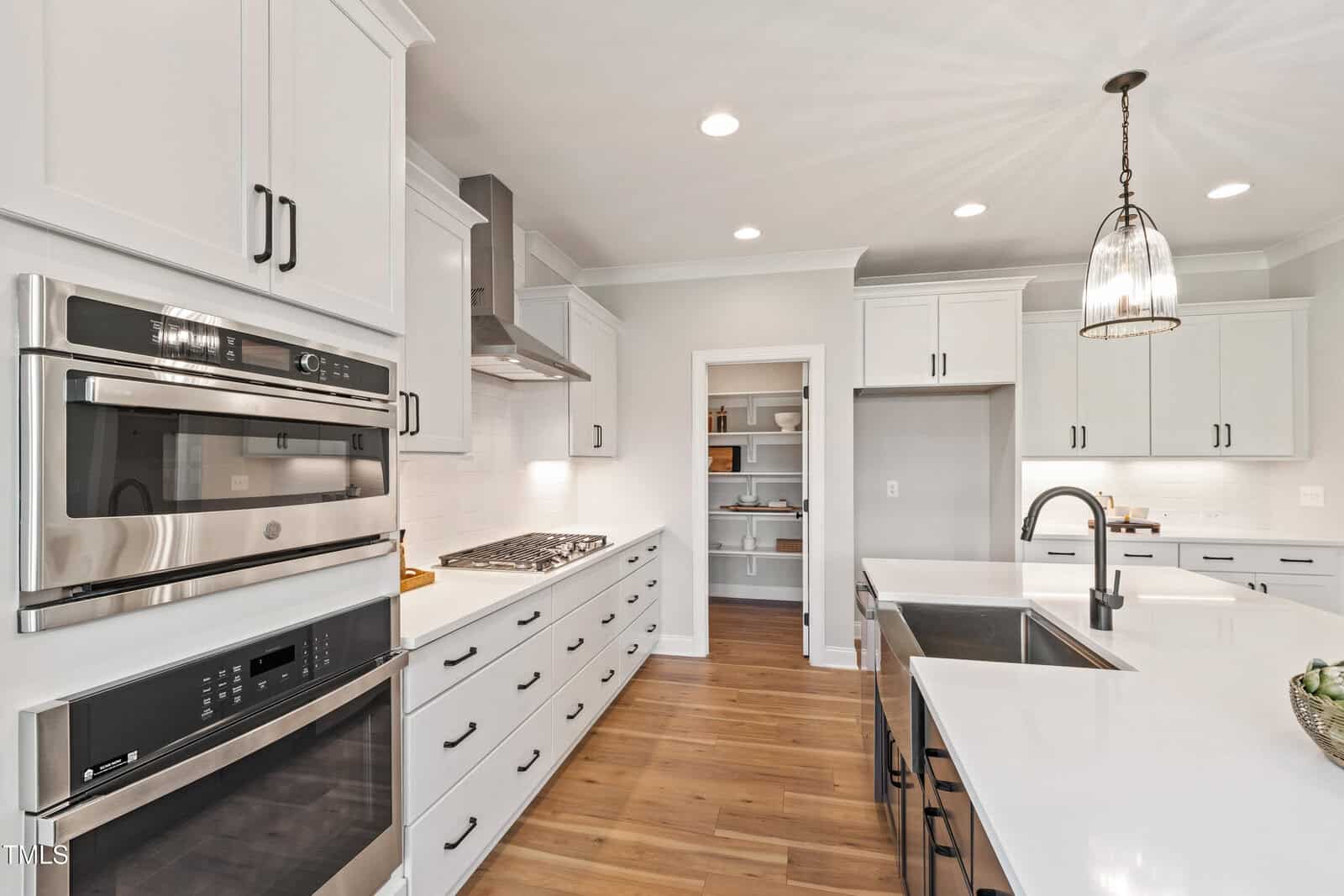Modern kitchen with white cabinets, stainless steel appliances, a center island with a sink, pendant lighting, wood flooring, and an open pantry in the back wall. The countertops are white and clutter-free.