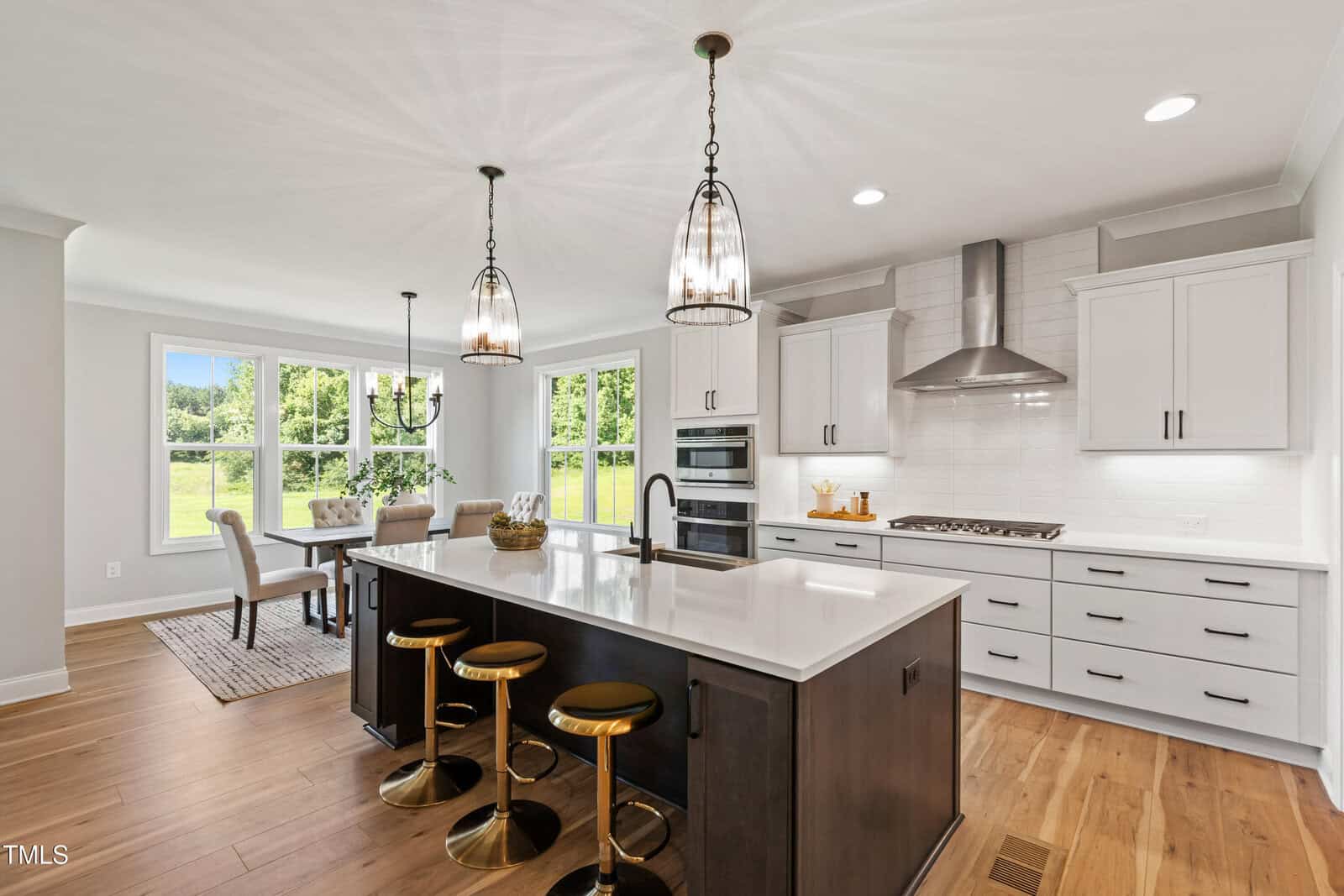 Modern kitchen with a large island, three gold and black barstools, white countertops, hanging pendant lights, white cabinets, stainless steel appliances, wood floors, and an adjacent dining area with large windows and a scenic outdoor view.