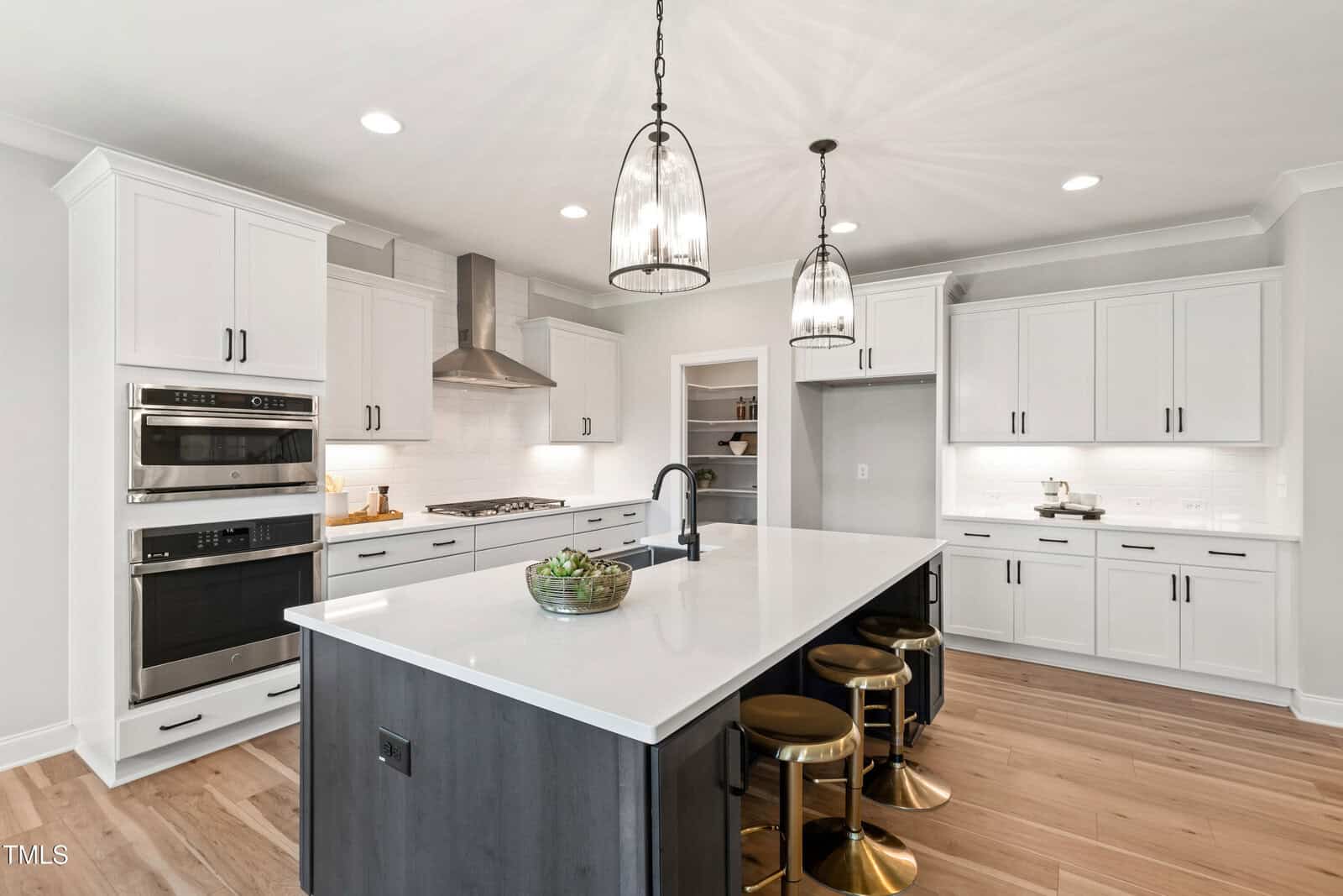 Modern kitchen with white cabinets, stainless steel appliances, a large island with a sink, gold stools, wood floors, and two pendant lights. Bowl of green apples sits on the island. Walls and backsplash are white.