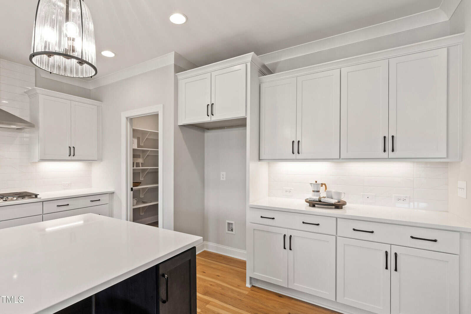 Modern kitchen with white cabinets, stainless steel hardware, light wood flooring, white tile backsplash, and a center island with a dark base. A pantry with open shelving is visible through a doorway.