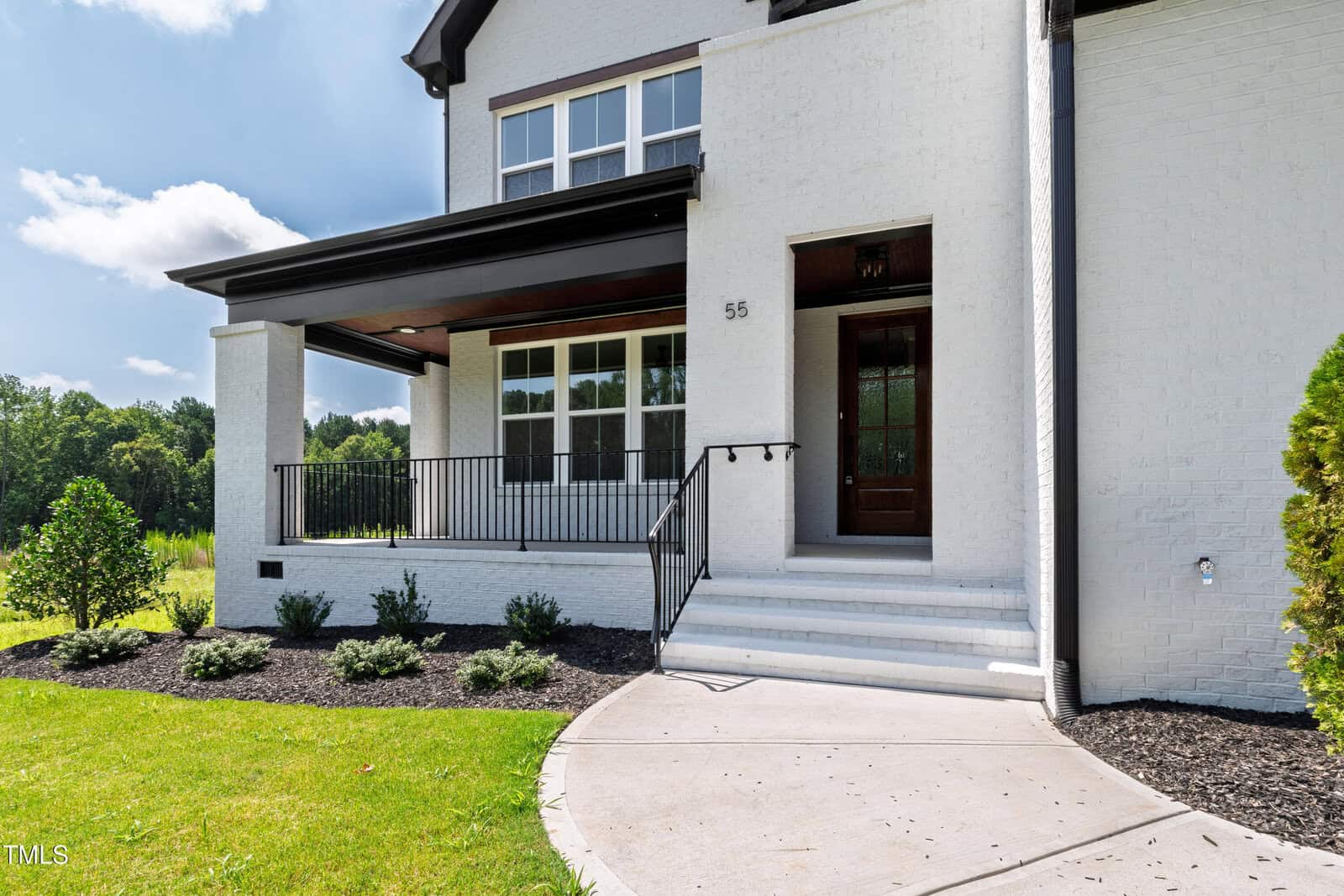 A modern white brick house with a dark-framed covered porch, black railing, wooden front door, and a curved concrete walkway. Green lawn and small shrubs line the front of the house.