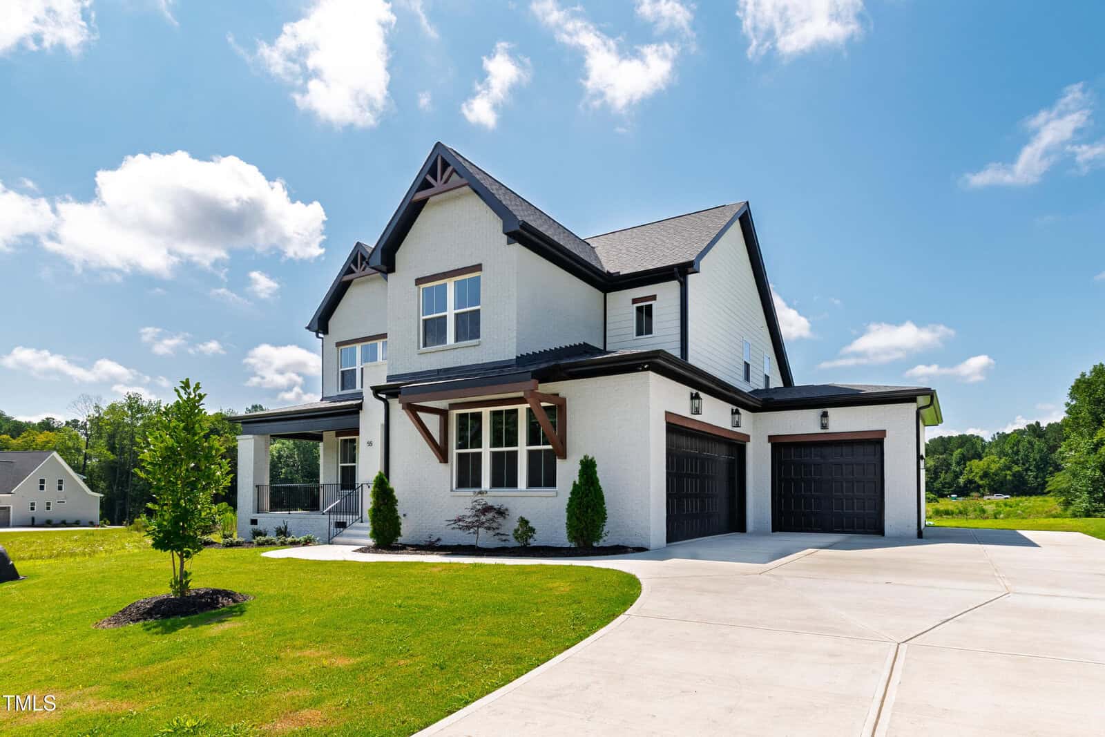 Two-story modern house with white exterior, dark trim, three-car garage, and a large driveway. The lawn is neatly landscaped, and the sky is mostly clear with a few scattered clouds. Trees and another house are visible in the background.