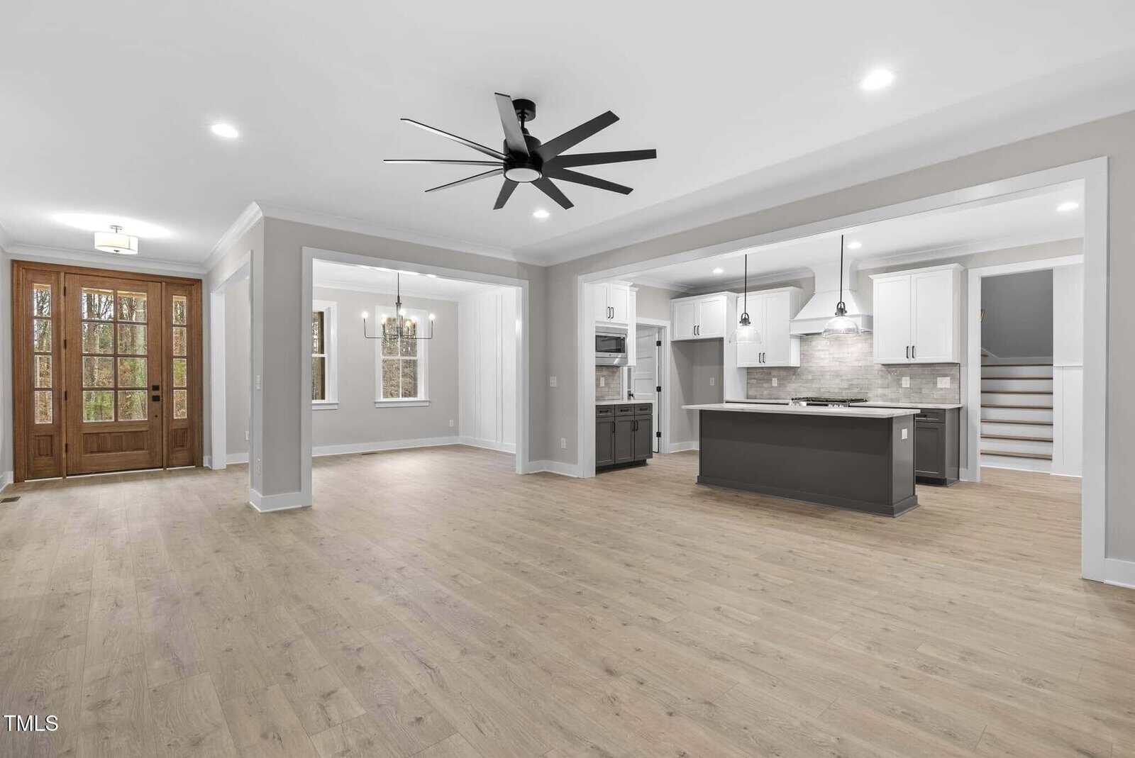 Spacious open-concept living area featuring a modern kitchen with an island, white cabinets, and stainless steel appliances. The room includes wood flooring, a large ceiling fan, and double wooden entry doors. Dining area and staircase in the background.