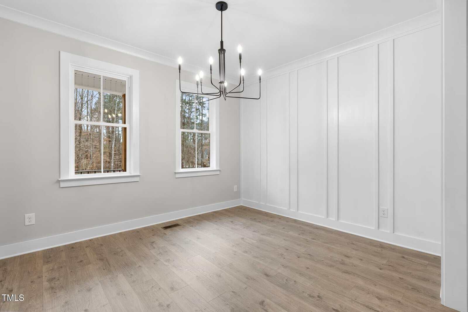 A bright, empty room with light wood flooring and light gray walls. Two windows on the left let in natural light. A modern black chandelier hangs from the ceiling. The right wall features decorative white paneling.