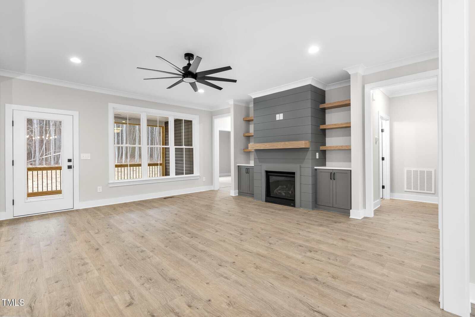 A modern living room featuring light hardwood floors, a ceiling fan, and a fireplace with gray paneling and floating shelves. Large windows and a glass door provide ample natural light. Walls are painted in a light neutral color.