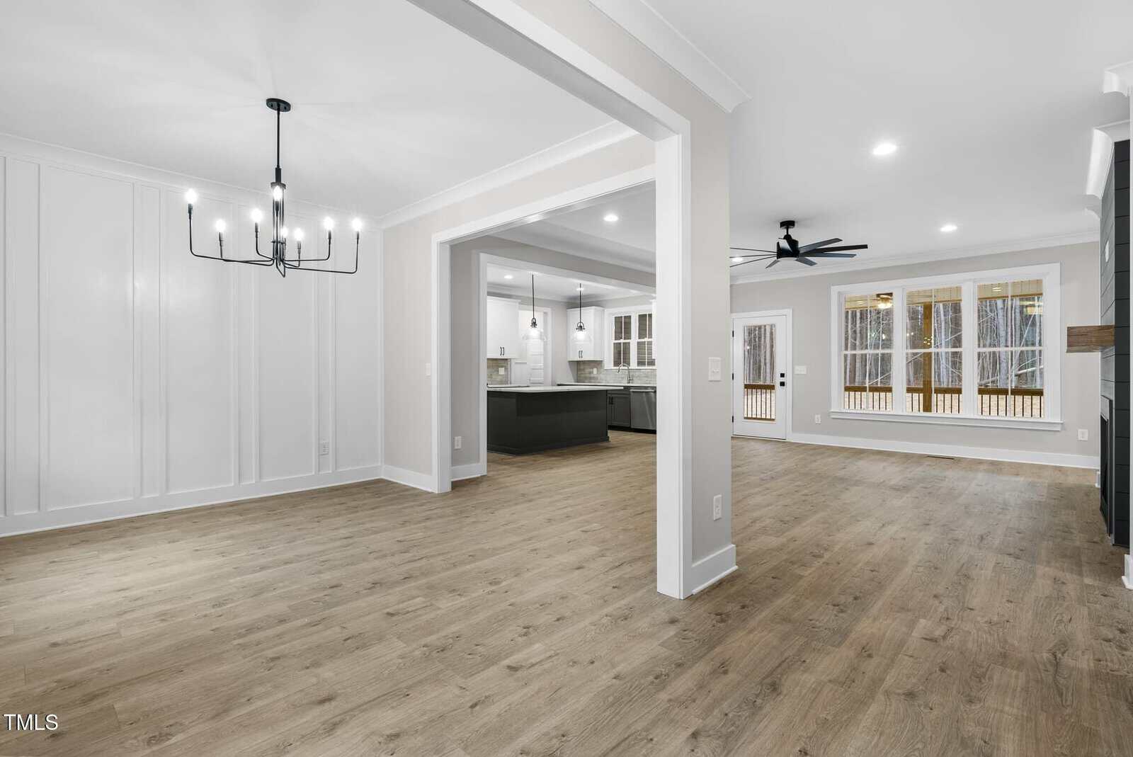 Open-concept interior with wooden flooring features a dining area with a modern chandelier, an adjacent living space with ceiling fan, and a view into a kitchen with light cabinetry. Large windows provide natural light.