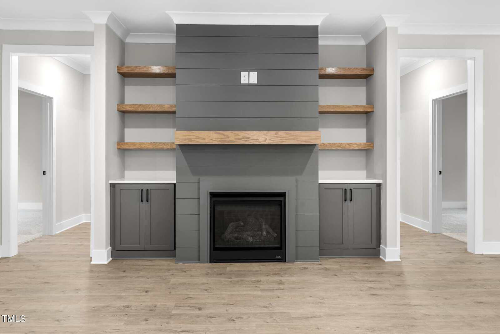 A modern living room features a central fireplace with gray shiplap paneling. There are built-in shelves and cabinets on either side. The room has light-colored walls, white trim, and wood flooring. Two doorways are visible in the background.