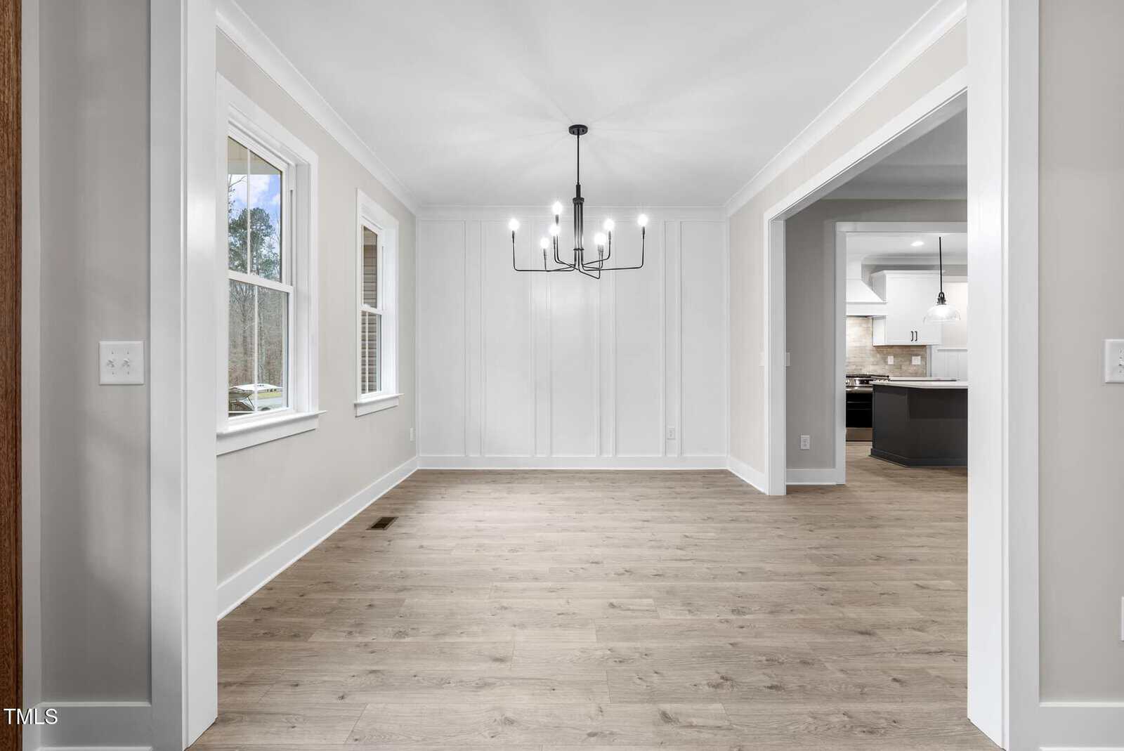 A spacious, empty dining room with light wood flooring and white walls. A black chandelier hangs from the ceiling. Large windows provide natural light. The room opens into a kitchen with visible countertops and cabinets.