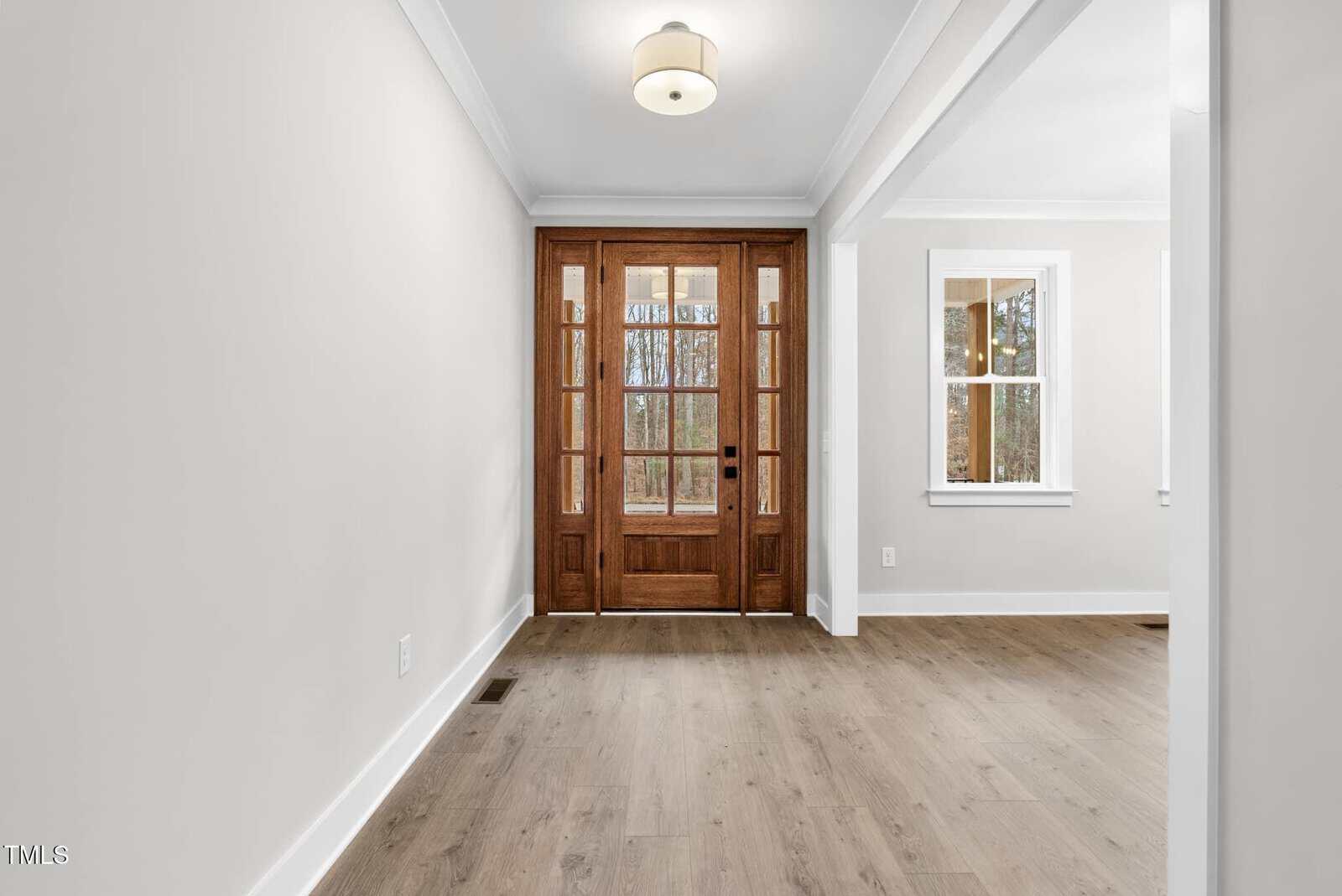 A bright entryway with light wood flooring features a wooden door with glass panels. The walls are painted cream, and a decorative light fixture is mounted on the ceiling. A window in the adjoining room allows natural light into the space.