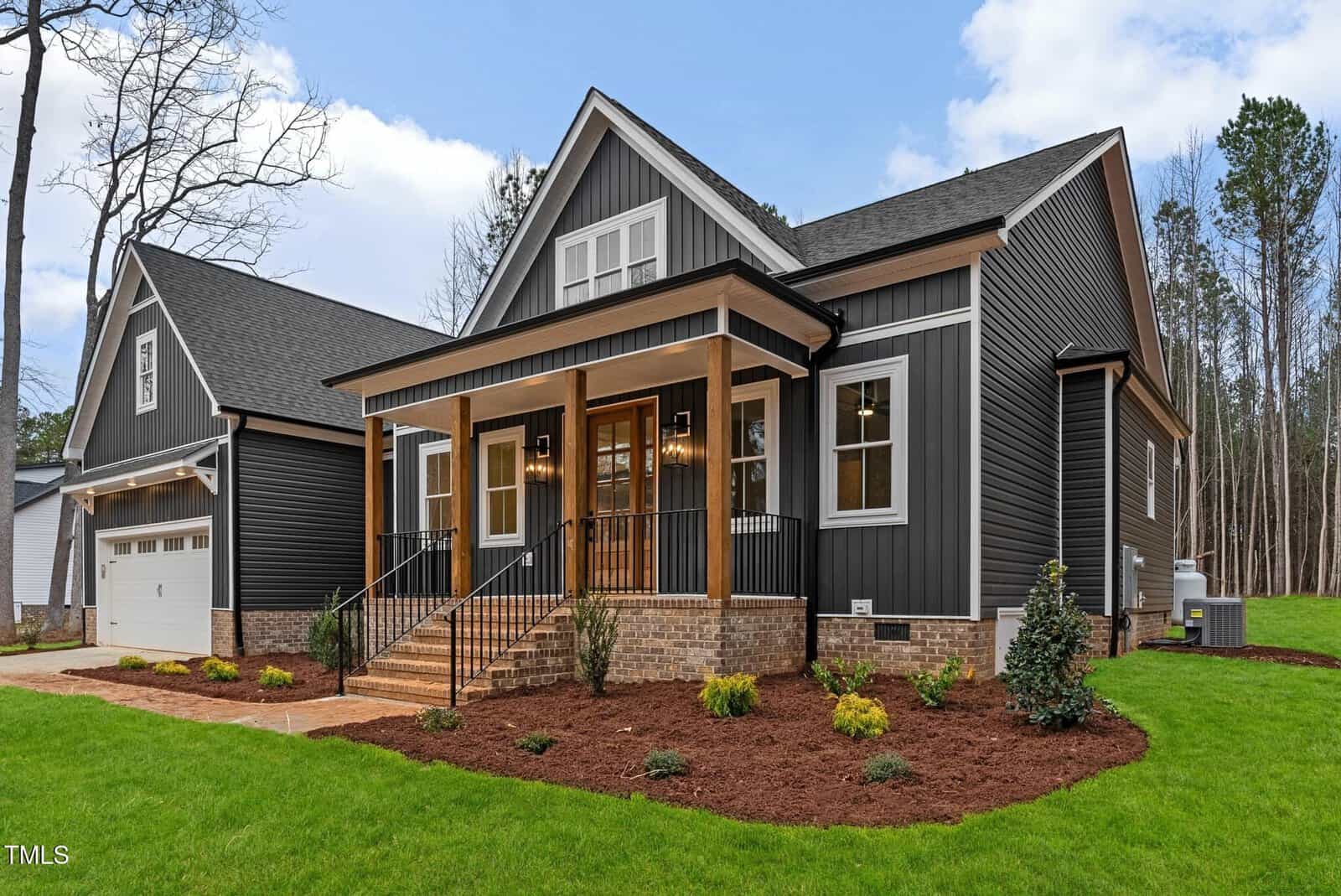 A modern black and white house with a wooden front porch and steps. The house features a two-car garage on the left and landscaped front yard with green grass and small shrubs. The background shows a row of tall trees under a blue sky.
