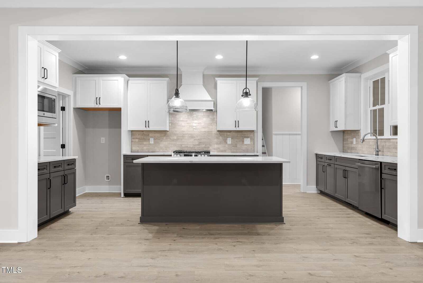 Modern kitchen with a large island featuring dark cabinetry, white upper cabinets, and pendant lighting. The room has a tiled backsplash, stainless steel appliances, and hardwood flooring.