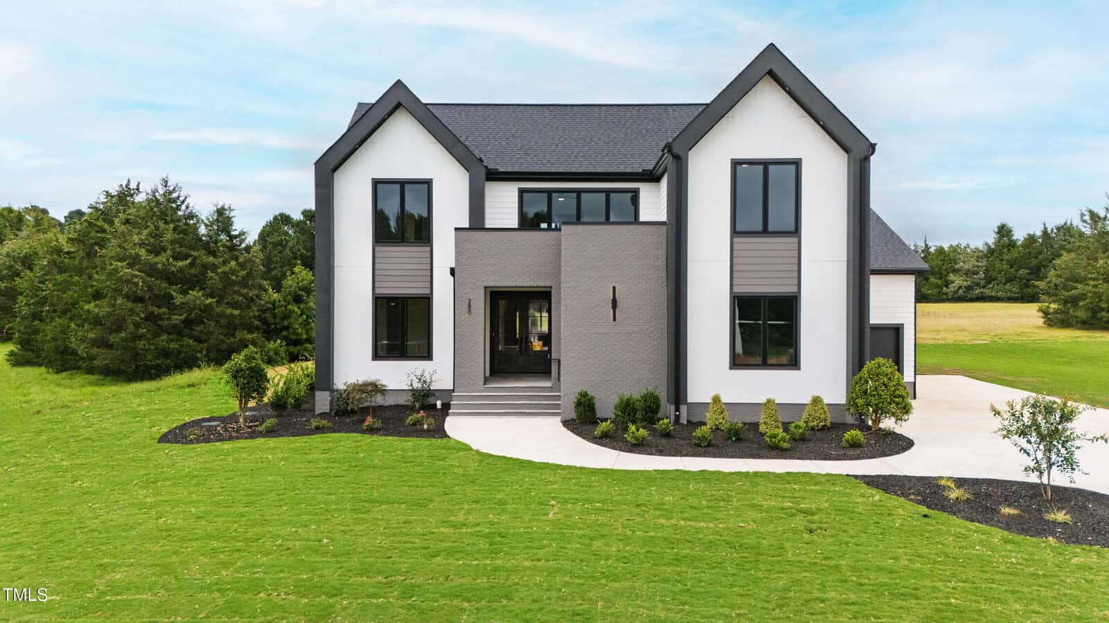 A modern two-story house with light gray walls, black trim, large windows, and a dark roof sits on a neatly landscaped lawn with shrubs, small trees, and a curved driveway at 280 Forest Bridge Road in Youngsville.