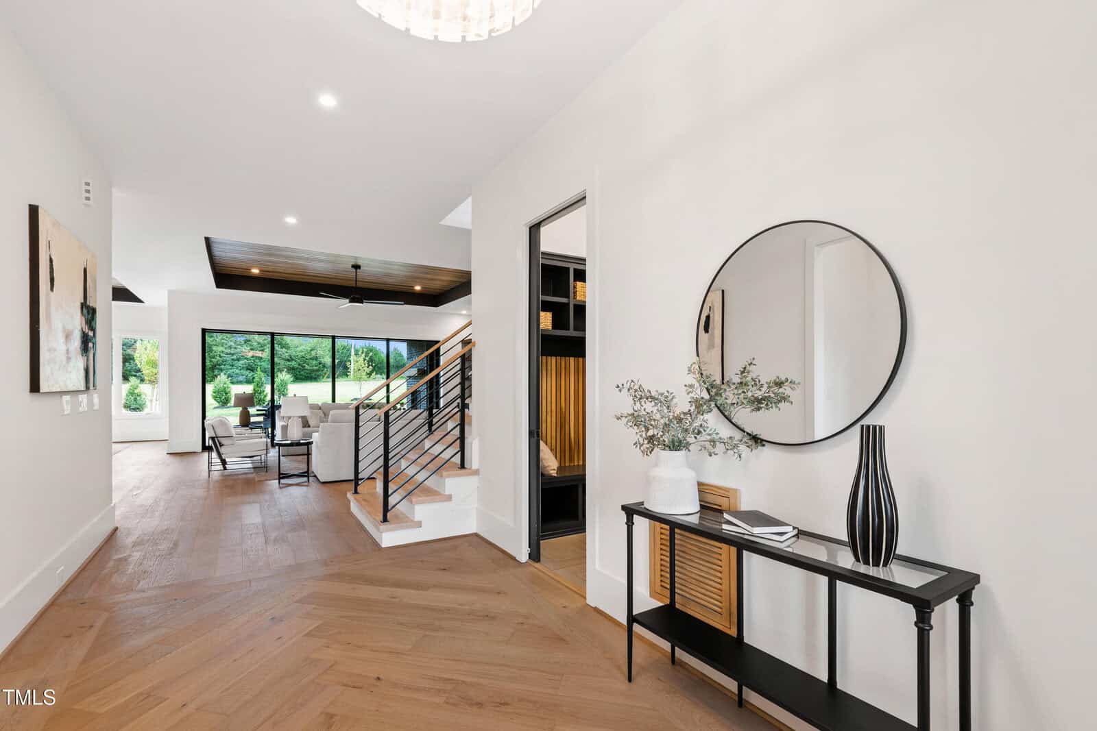 A modern entryway with light wood floors, a black console table, a round mirror, decorative vases, and a staircase with metal railings. The space opens to a living area with large windows and greenery outside.