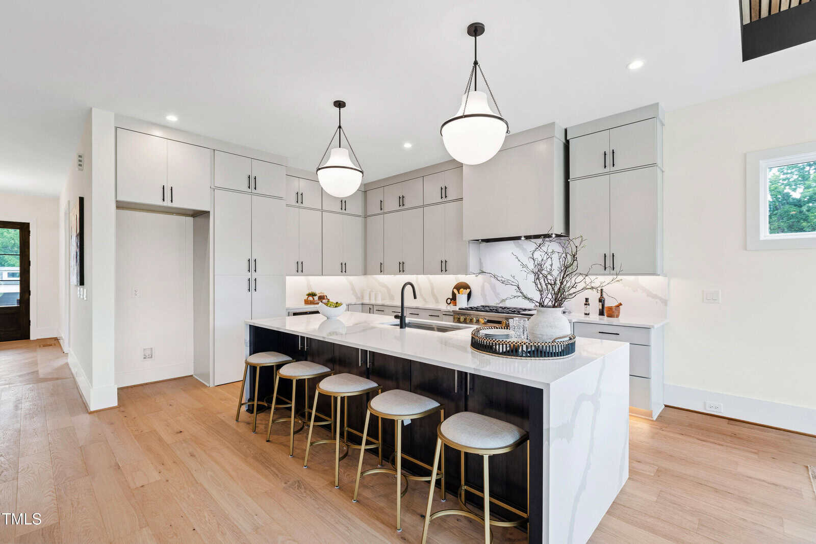 Modern kitchen with white cabinets, black accents, a large island with four gold stools, pendant lights, wood flooring, and a decorative tray with branches on the countertop.