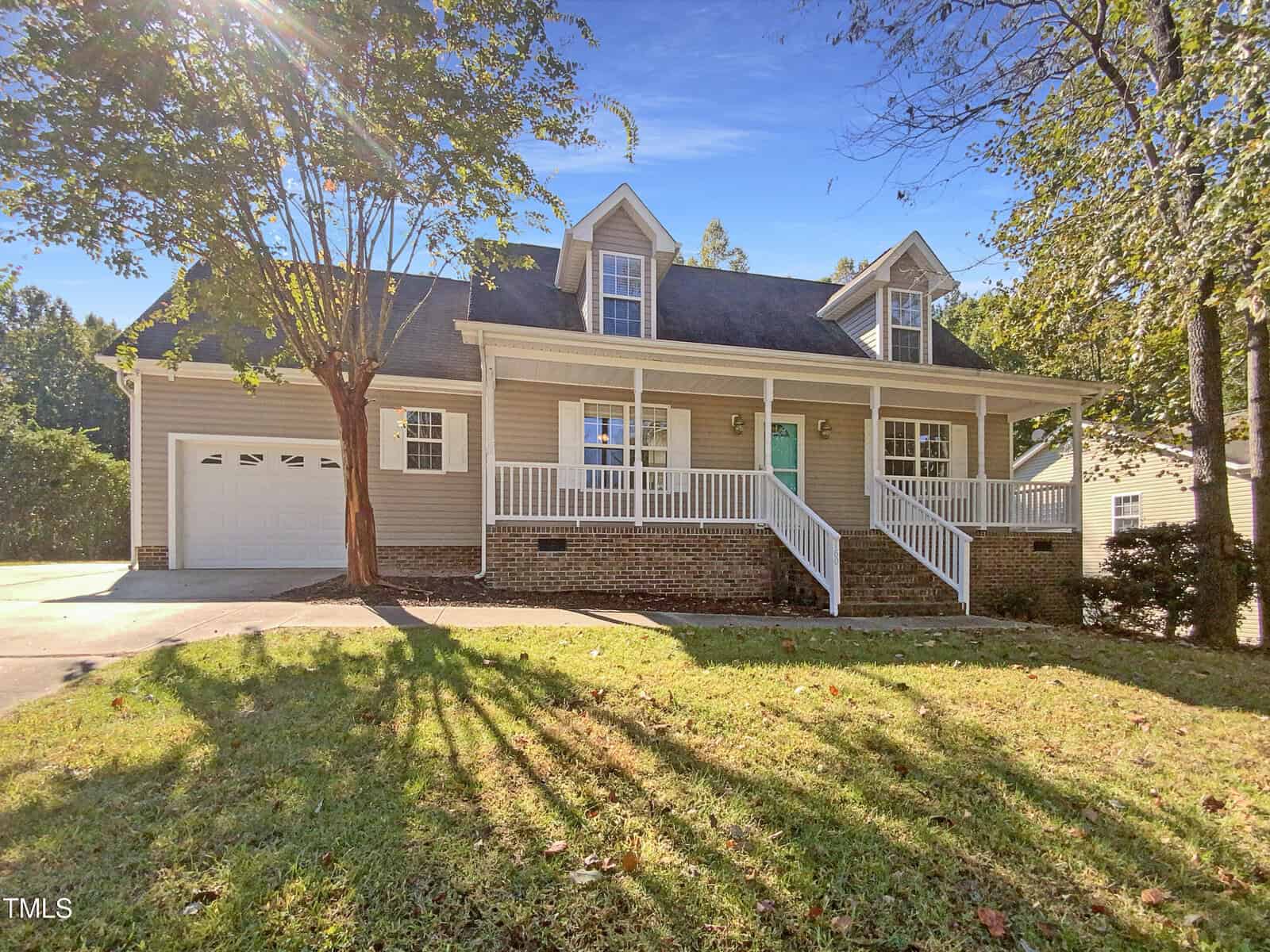 A two-story house with beige siding, brick foundation, white trim, and a covered front porch. Two dormer windows are on the roof. There is an attached two-car garage and a sloped front yard with grass and trees.