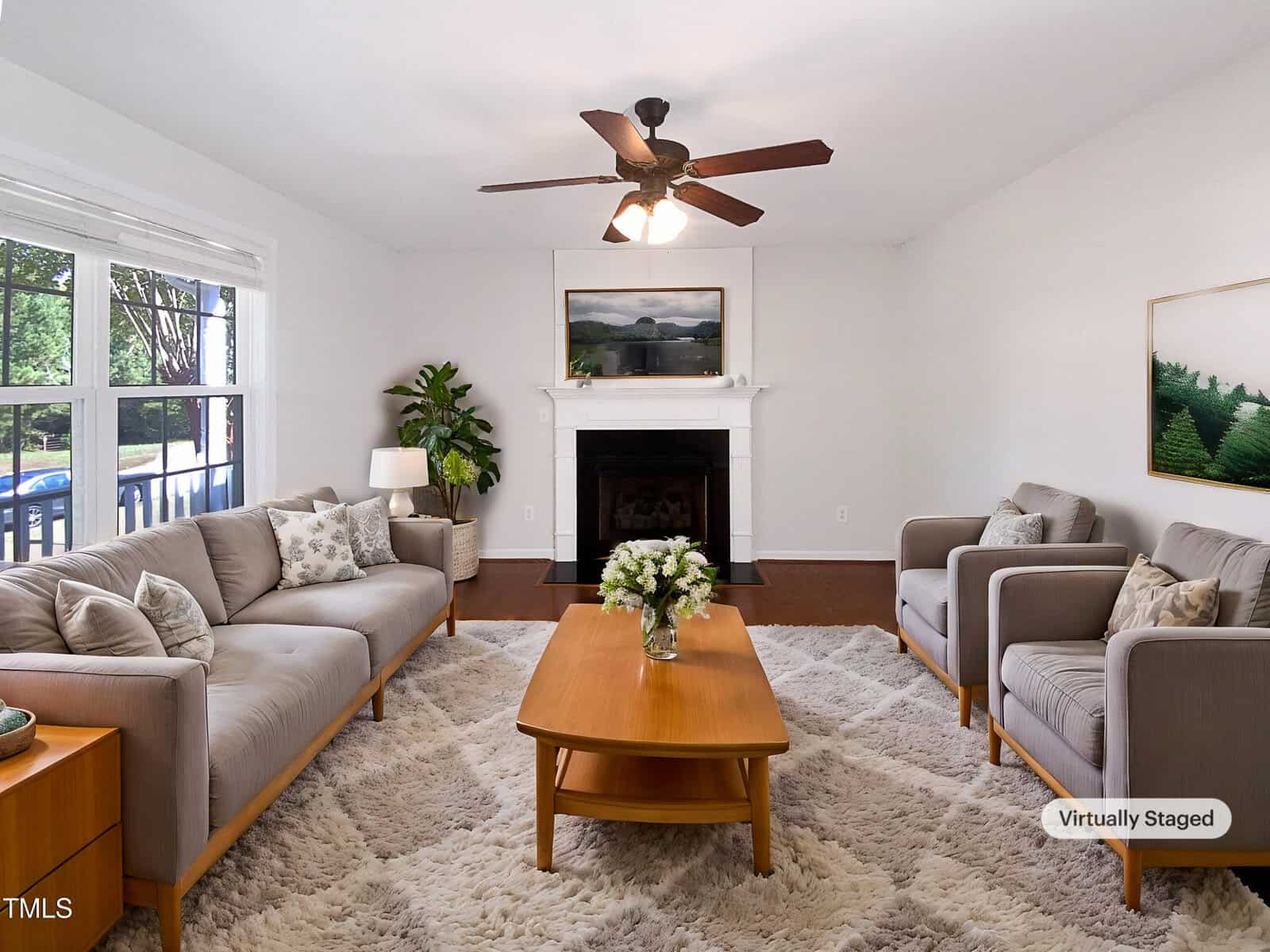 A living room with two armchairs and a sofa, a wooden coffee table on a light shag rug, a ceiling fan, a fireplace with art above it, and large windows showing trees outside. A "Virtually Staged" label is visible in the corner.