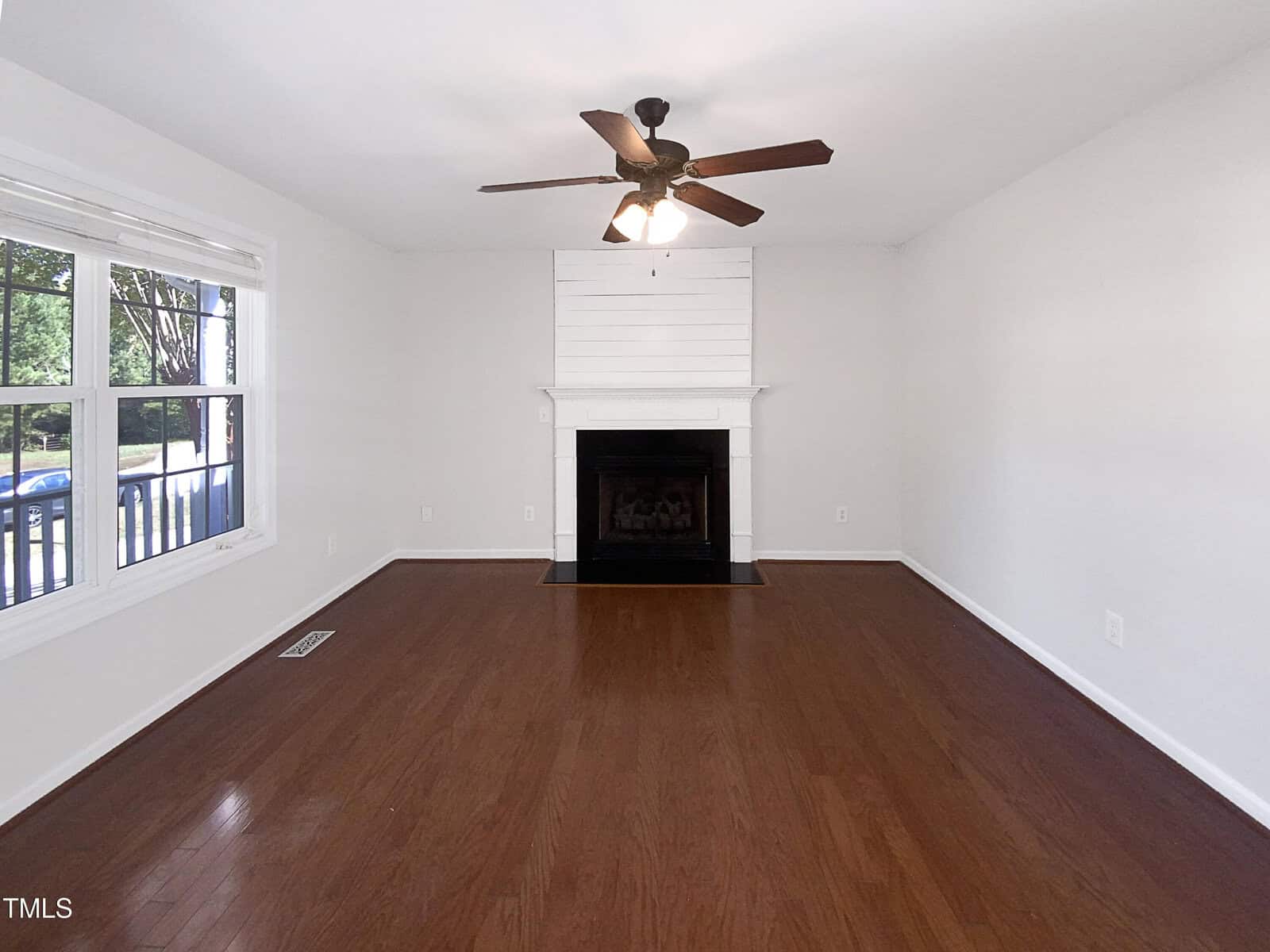 A living room with hardwood floors, white walls, a black fireplace with a white mantel, a ceiling fan with lights, and a large window showing trees and a front porch railing.