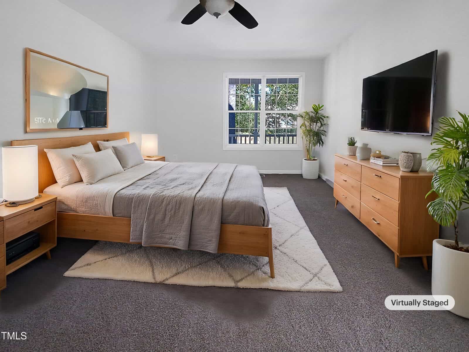 A bedroom with a large bed, nightstands with lamps, a dresser, potted plants, a wall-mounted TV, a ceiling fan, a window with a view of trees, and a neutral color palette. The image is virtually staged.