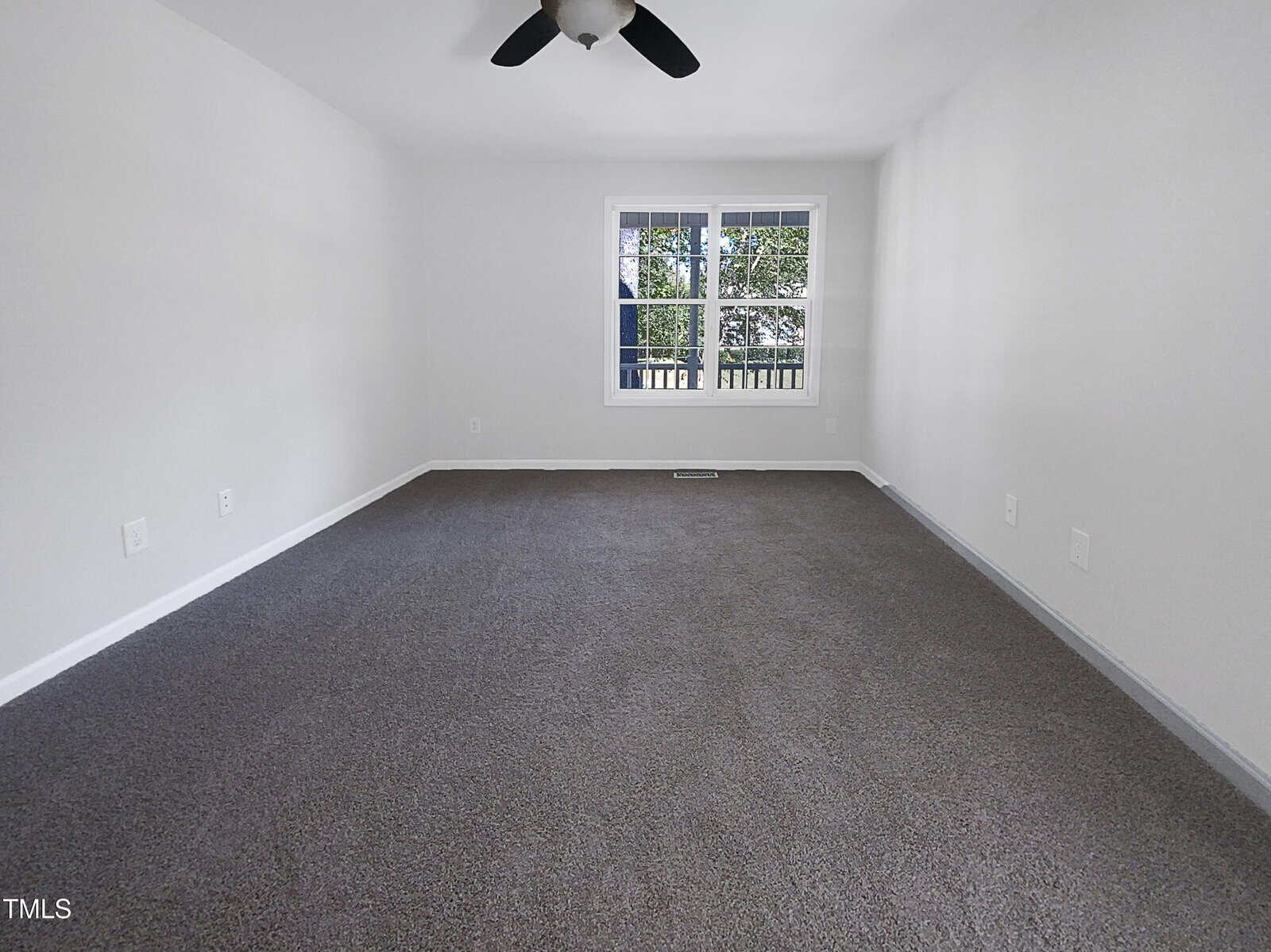 Empty room with white walls, gray carpet, a ceiling fan with light, and a window showing trees outside. The room appears bright with natural light and has electrical outlets along the walls.