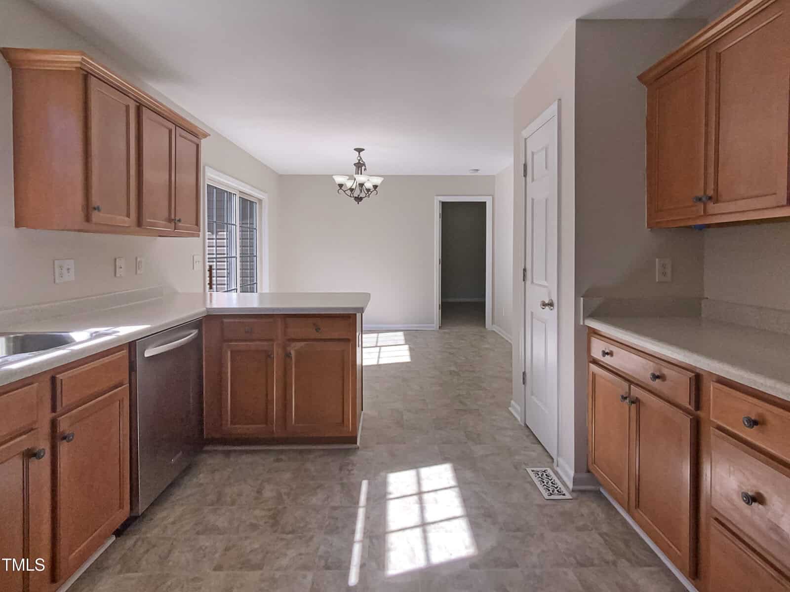 A kitchen with wood cabinets, light-colored countertops, stainless steel appliances, and tile flooring, with sunlight coming through a window. A dining area with a chandelier is visible in the background.