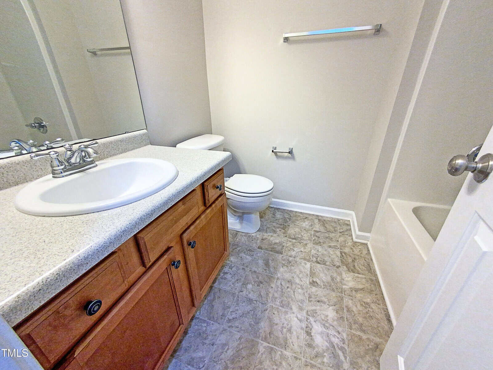 A bathroom with a white sink set in a speckled countertop, wooden vanity, toilet, bathtub, wall-mounted towel bar, toilet paper holder, and light-colored tile flooring.