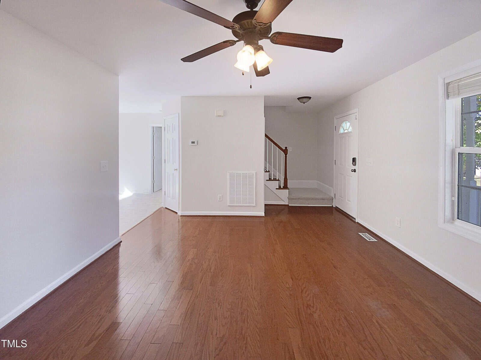 A bright, empty room with hardwood floors, white walls, a ceiling fan with lights, a window on the right, a staircase near the entryway, and a white front door with a small window.
