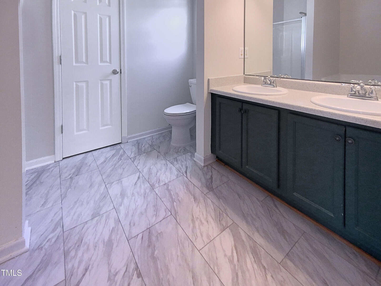 A bathroom with light gray marble tile flooring, a white toilet, a double-sink vanity with dark cabinets, a large mirror, and a closed white door. The walls are painted a light neutral color.