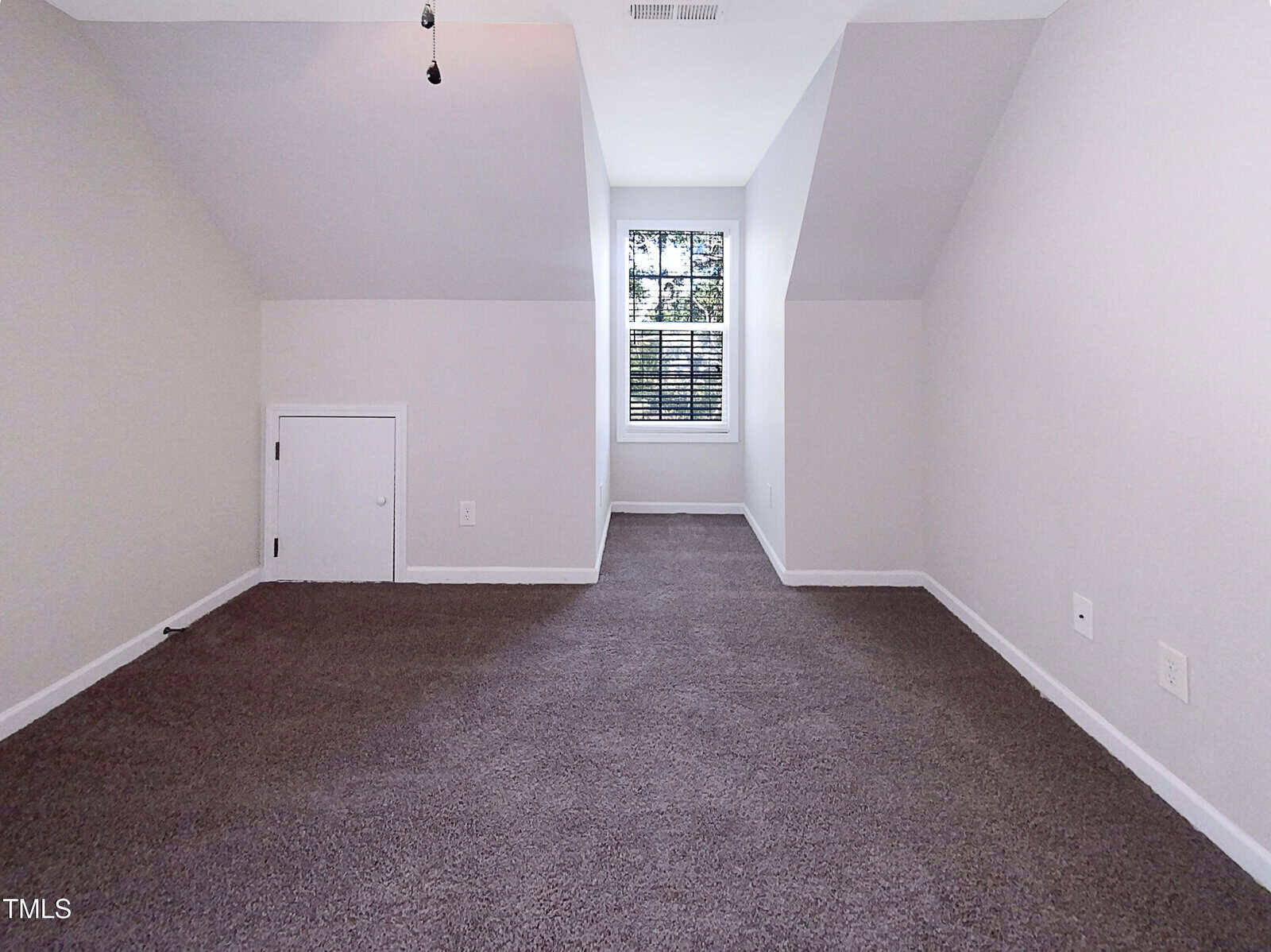Room with sloped ceilings and gray carpet, a small white door on the left wall, white walls, a central window with blinds, and two electrical outlets visible on the right wall. Ceiling light fixture lacks a bulb or cover.