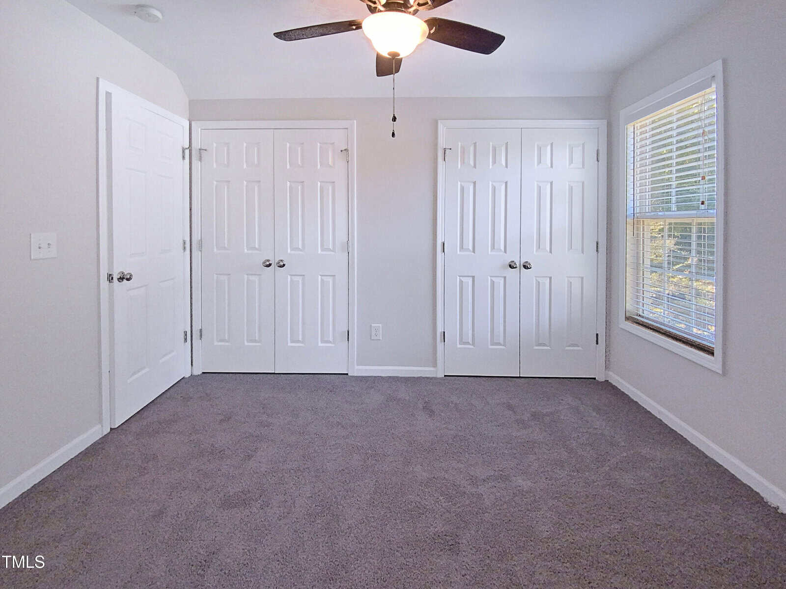 Bright, empty bedroom with gray carpet, white walls, ceiling fan, two double-door closets, and a large window with blinds letting in natural light.