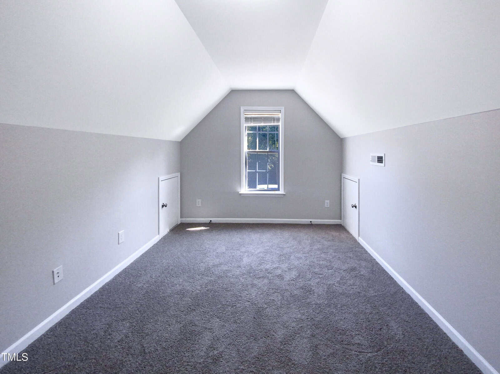 A bright, empty attic room with sloped white ceilings, gray carpet, a single window at the end, and two small closets built into the side walls.