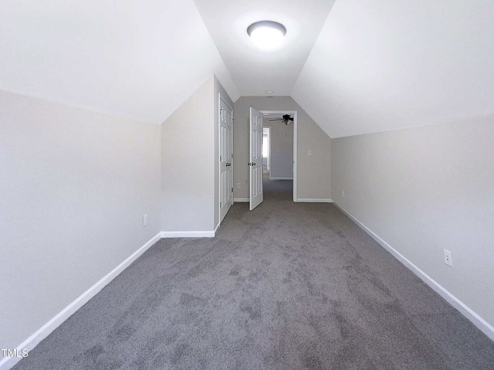 A long, narrow attic room with sloped white walls, gray carpet, recessed lighting, and an open door at the far end leading to another room.