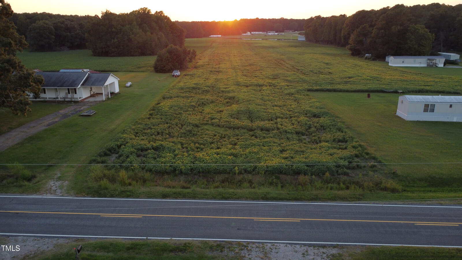 A grassy, rectangular field bordered by trees and mobile homes is shown at sunset, with a road in the foreground and a house on the left side. The sky is orange as the sun sets in the background.