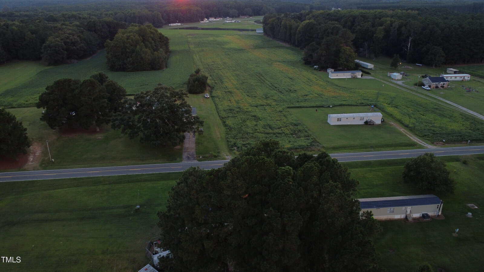 Aerial view of a rural area with green fields, scattered trees, a few houses, and a road running through the landscape at sunset. Dense forest borders the area in the background.