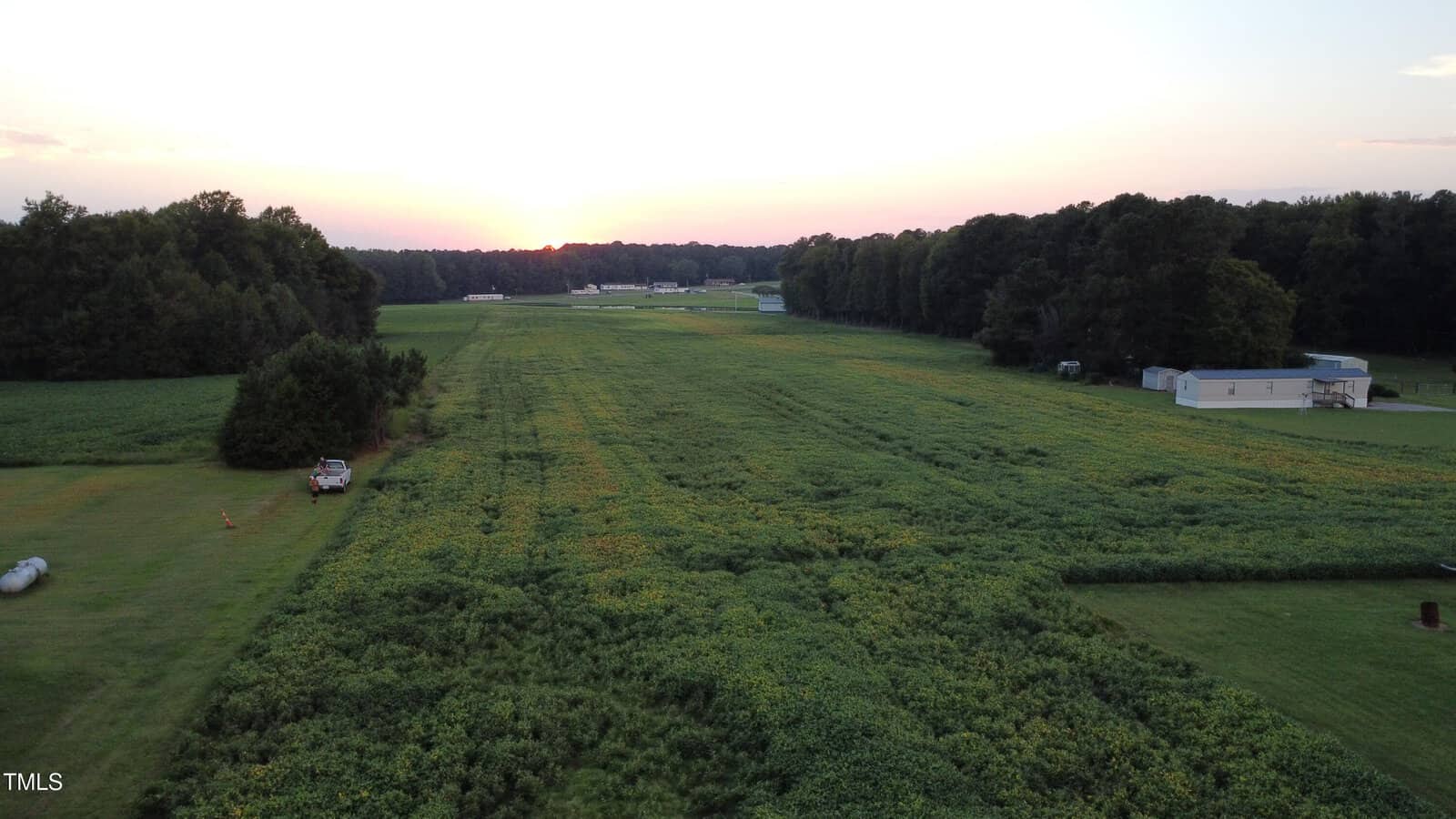 Aerial view of a rural landscape at sunset, showing a large green field bordered by trees, a parked vehicle, and several small buildings or mobile homes along the edge of the field.