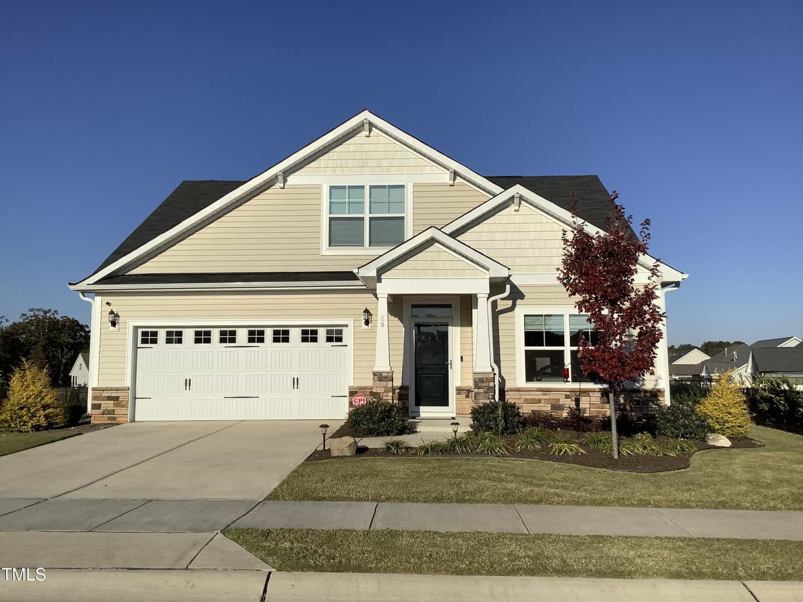 A modern two-story house with beige siding, white trim, and a front porch. There is a double garage with white doors, a concrete driveway, and a manicured lawn with a small red-leafed tree near the entrance.