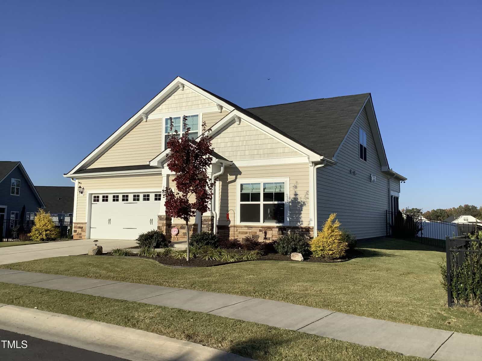 A beige single-family house with a two-car garage, white trim, and dark roof sits on a neatly manicured lawn with small trees and shrubs, under a clear blue sky.