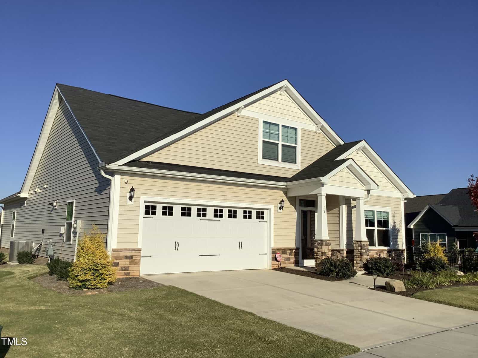 A beige single-story house with white trim and a dark roof, featuring a two-car garage, small front porch, and neatly landscaped yard on a sunny day.