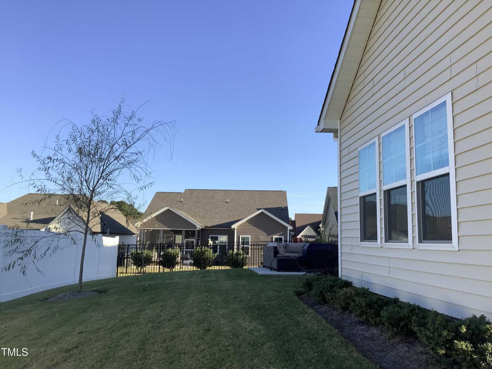 A yellow house with white trim and several windows is shown on the right, with a grassy backyard, a small tree, a white fence, and neighboring houses visible in the background under a clear blue sky.