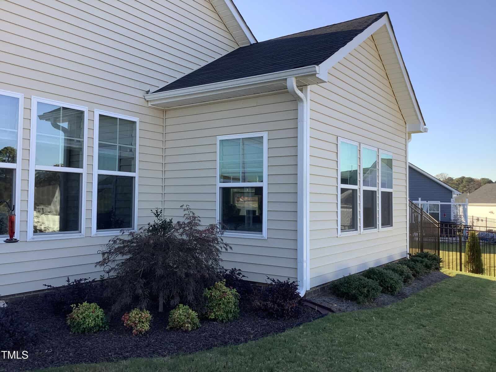 A beige house with white trim and multiple windows, light brown siding, and a black shingle roof. Neatly landscaped garden beds with shrubs and small bushes line the exterior. Grass and a black fence are visible.
