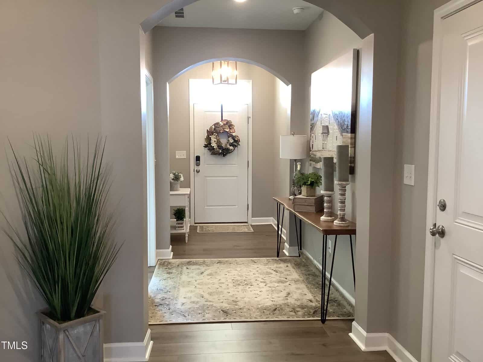 A hallway with wood floors, a rug, console table with decor, a large potted plant, and a white front door decorated with a floral wreath under a modern light fixture.
