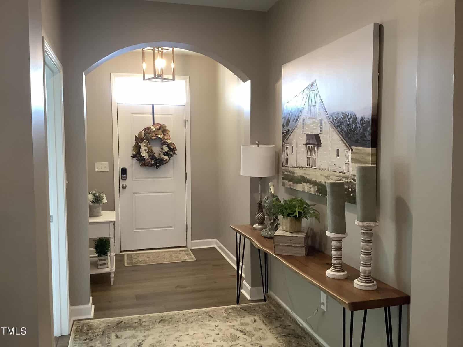 A hallway with a door decorated with a wreath, a patterned rug, a console table with lamps, plants, and candles, and a large farmhouse painting on the wall. Light gray walls and wood floors create a cozy ambiance.