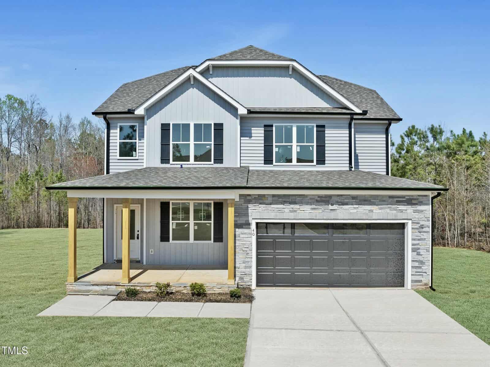 A two-story modern house with light gray siding, black shutters, a covered front porch with wooden pillars, a double garage, stone accents, and a well-kept lawn under a clear blue sky.