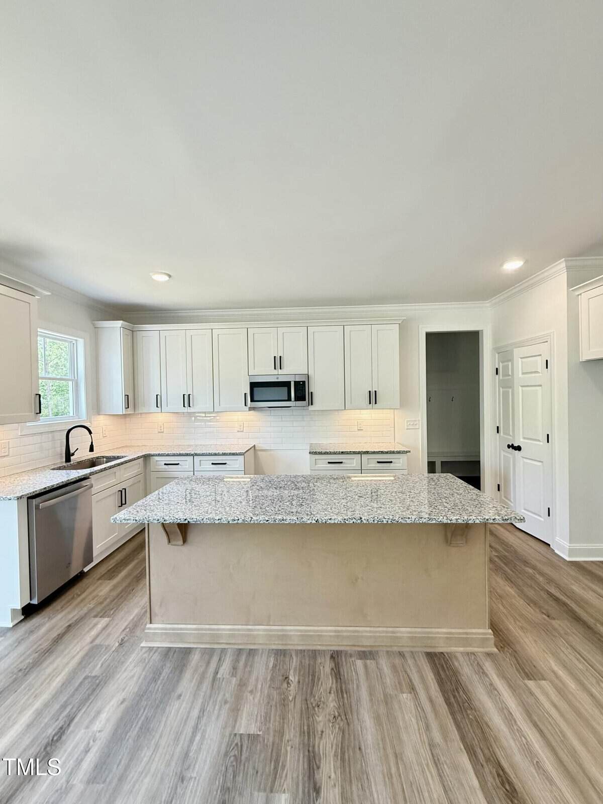 A modern kitchen with white cabinets, stainless steel appliances, a granite island countertop, subway tile backsplash, and light wood flooring. The space is well-lit with recessed lighting and natural light from a window.