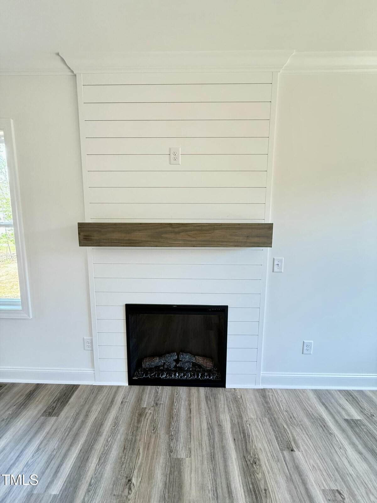 A modern fireplace with a dark wood mantel and white shiplap wall, set in a room with light-colored wood flooring and white walls. A small outlet or switch is above the mantel.