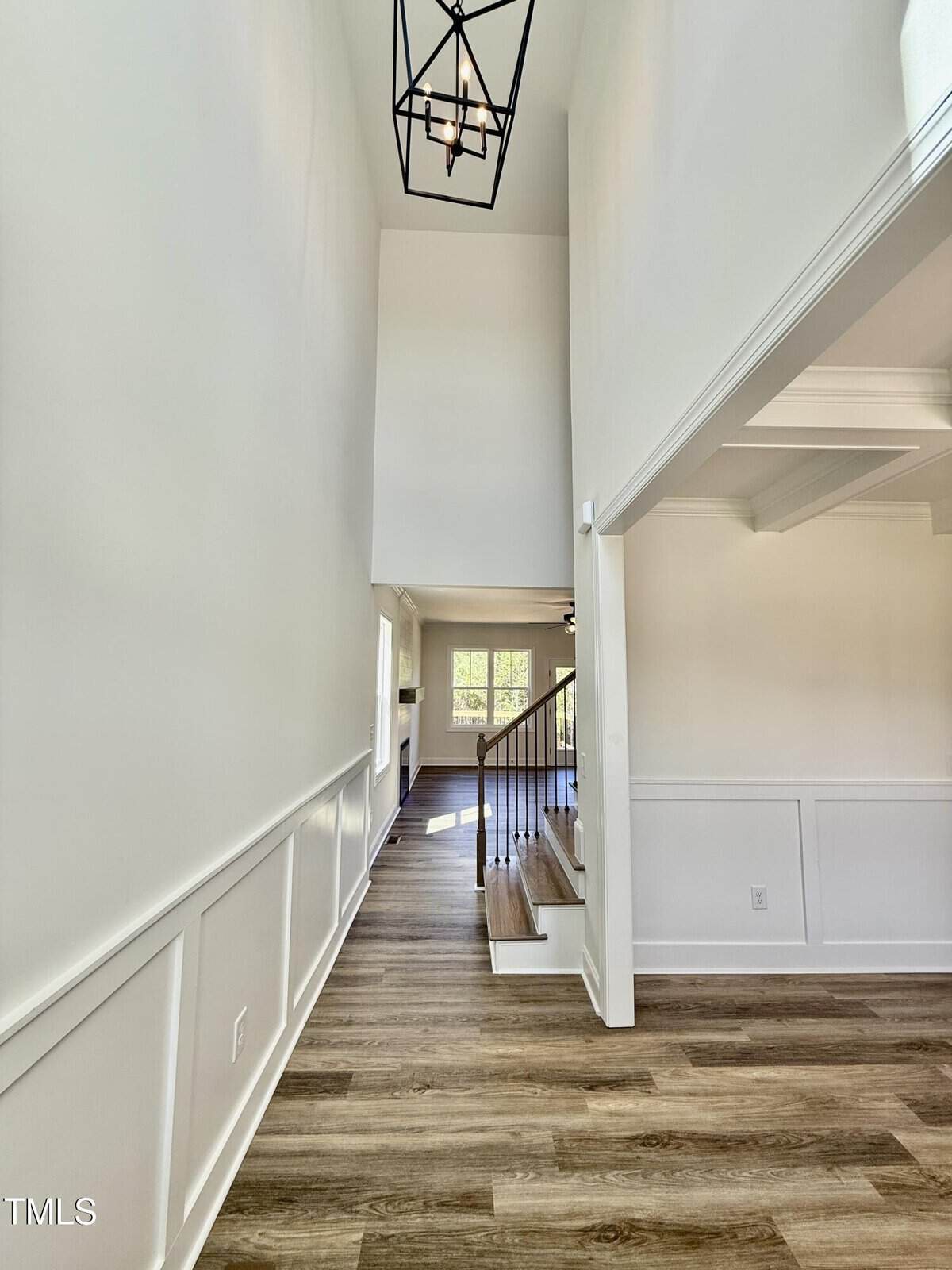 Entry hallway of a modern home with light-colored walls, wood flooring, white wainscoting, a staircase with a wooden handrail, and a geometric chandelier overhead. Natural light enters from windows in the back.