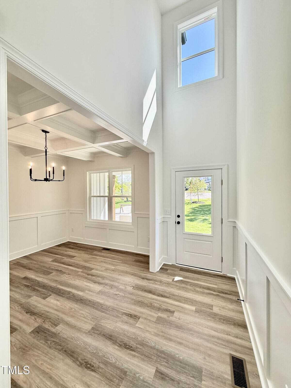 A bright entryway with high ceilings, white walls, large windows, a glass-paneled front door, and wood-look flooring. An adjacent dining area features a black chandelier and coffered ceiling.