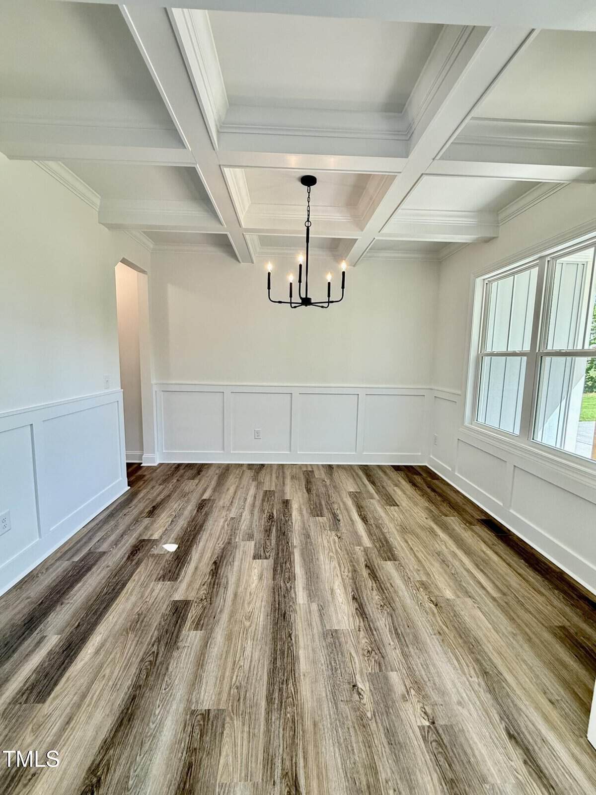 A dining room with white coffered ceiling, white wainscoting, wood-look flooring, a black chandelier, large windows on the right, and an open doorway on the left. The room is empty.
