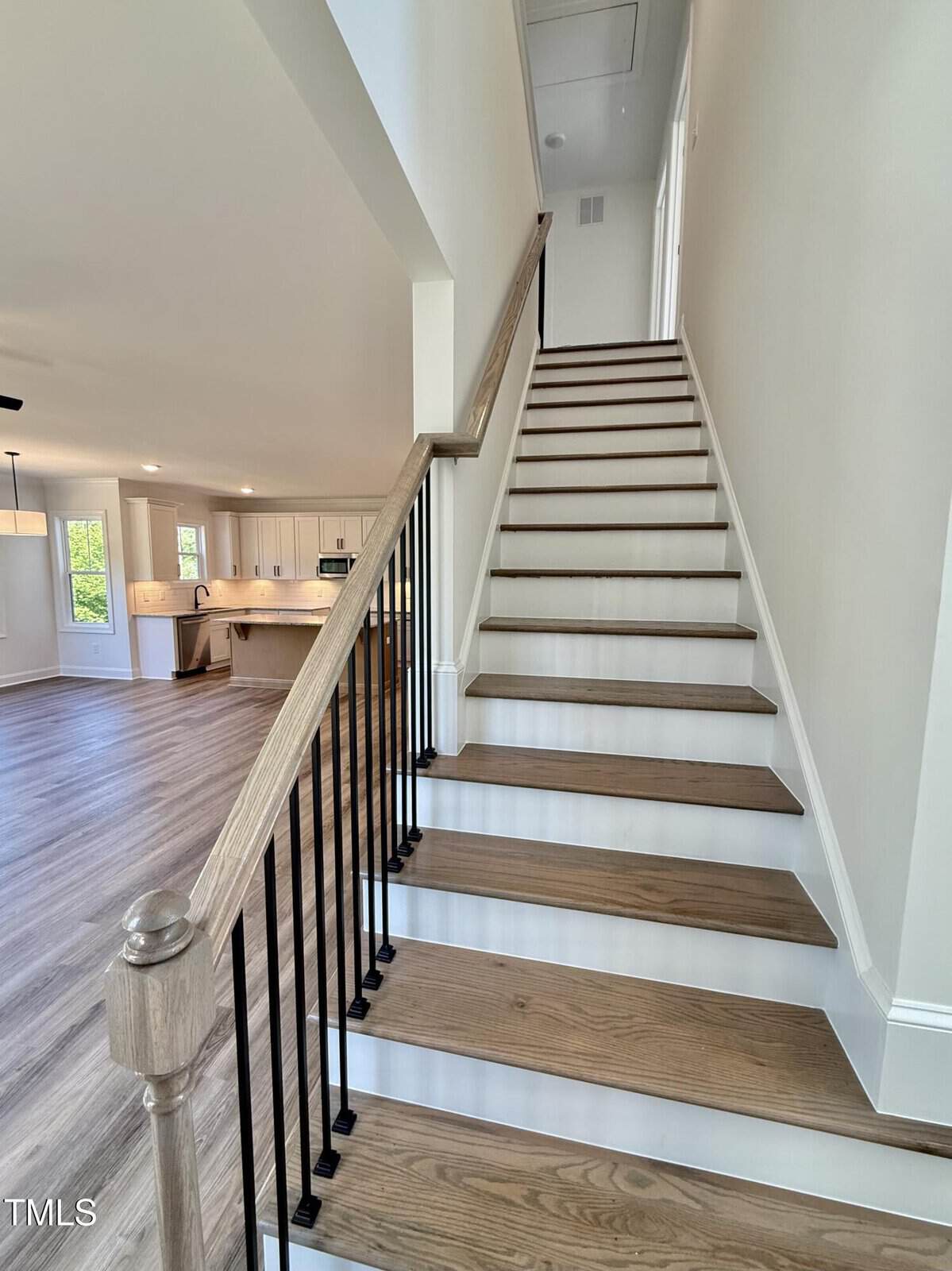 A wooden staircase with black balusters leads to an upper floor in a modern home. The stairs are adjacent to an open-concept kitchen and living area with light wood flooring and white walls.