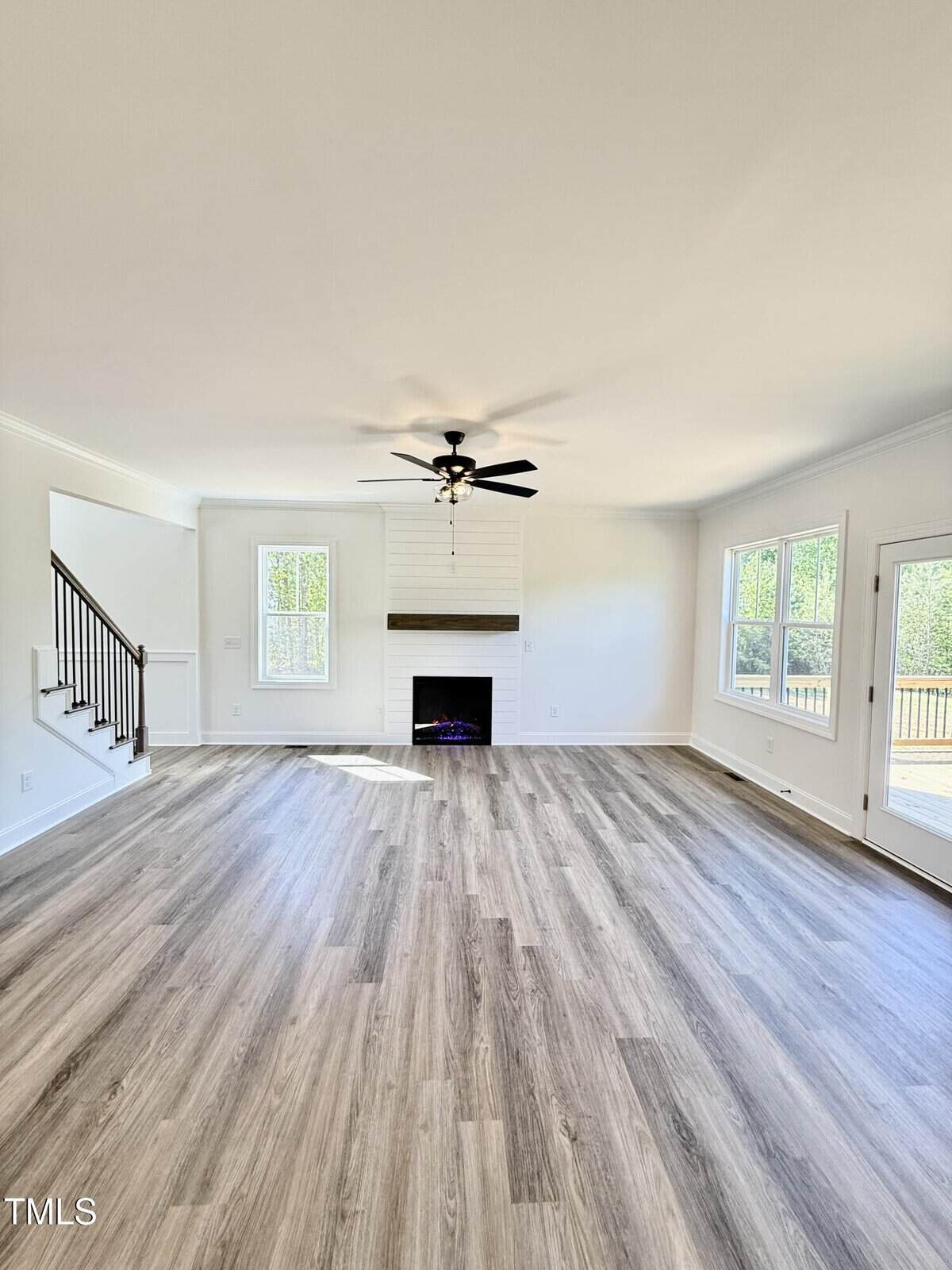 A spacious, empty living room with light wood flooring, white walls, a ceiling fan, a fireplace centered on one wall, large windows, and a door leading to an outdoor deck. Stairs are visible to the left.