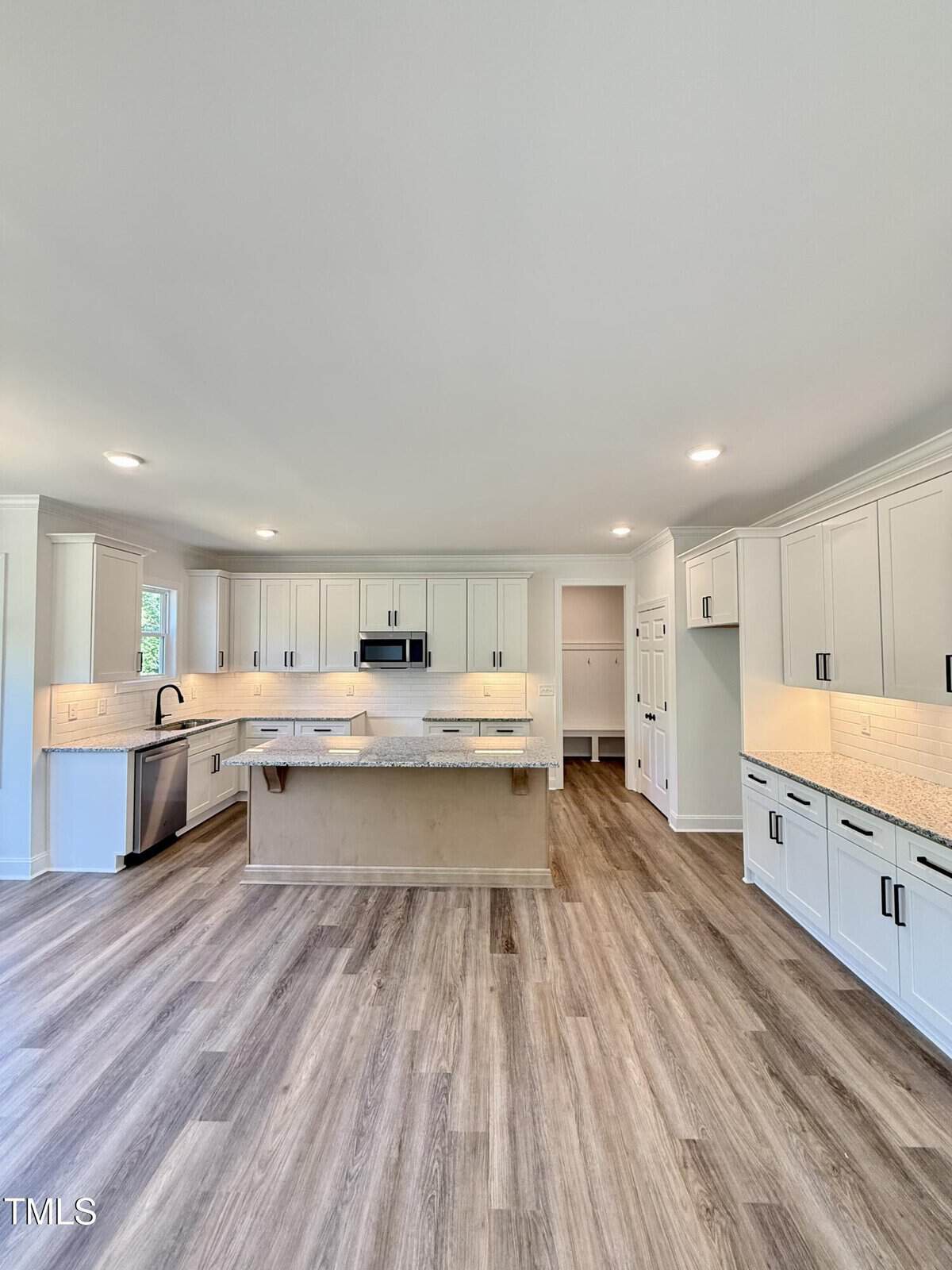 A spacious kitchen with white cabinets, granite countertops, a central island, stainless steel appliances, under-cabinet lighting, and wood-look flooring. The room is well-lit with recessed ceiling lights.